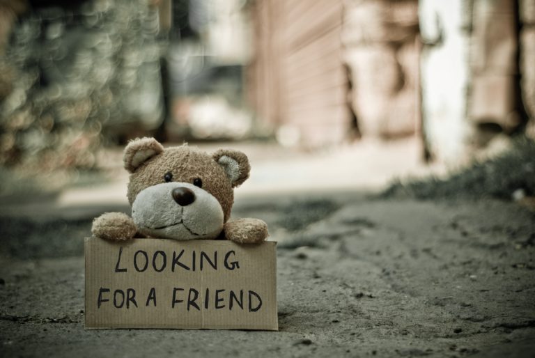 Petit ourson en peluche tenant une pancarte où il est inscrit "Looking for a friend" / En recherche d'amis